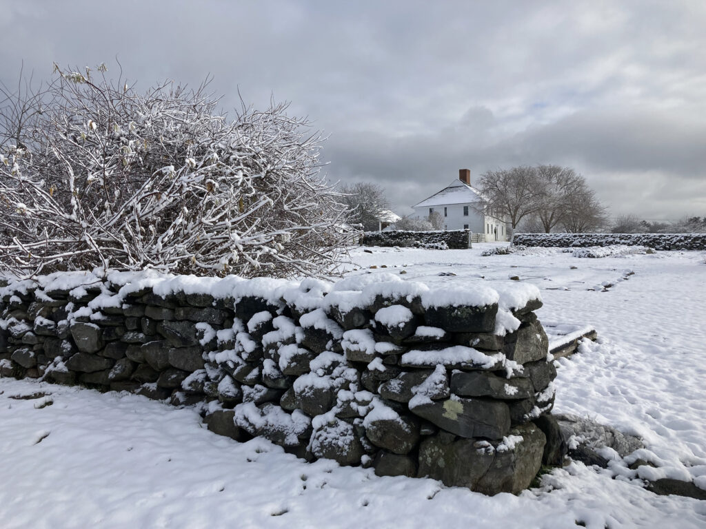 Snow-covered stone wall and bush with a distant farmhouse and leafless trees under a cloudy sky.