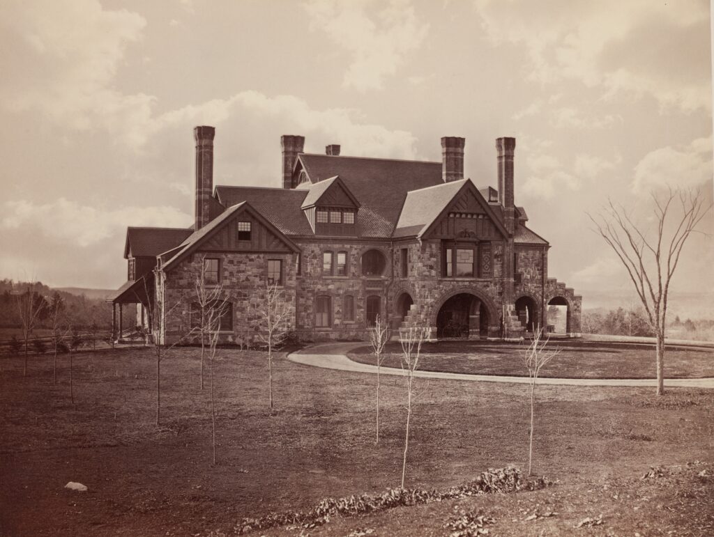 Historic stone mansion with tall chimneys surrounded by barren trees and expansive lawn under a cloudy sky.