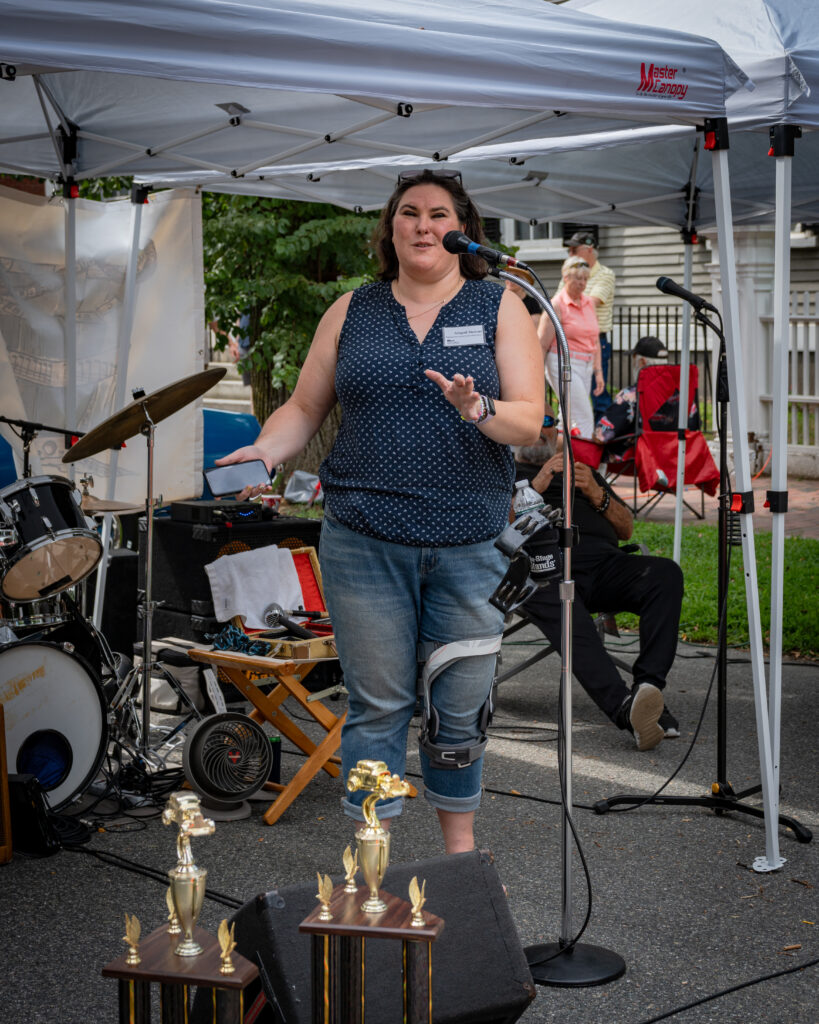 A woman speaks into a microphone at an outdoor event under a tent, with musical instruments and trophies nearby.