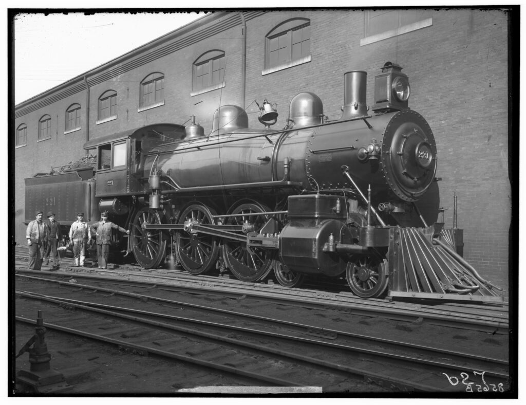 Vintage steam locomotive with five men beside it, in front of a brick industrial building with arched windows.